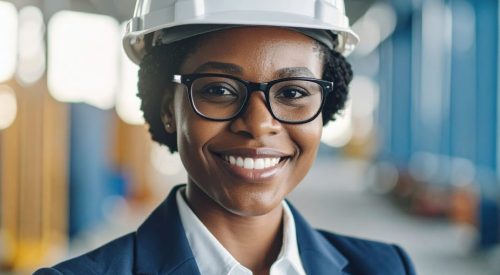 A confident female engineer wearing hard hat and glasses smiles warmly in construction environment, showcasing her professionalism and expertise in industry